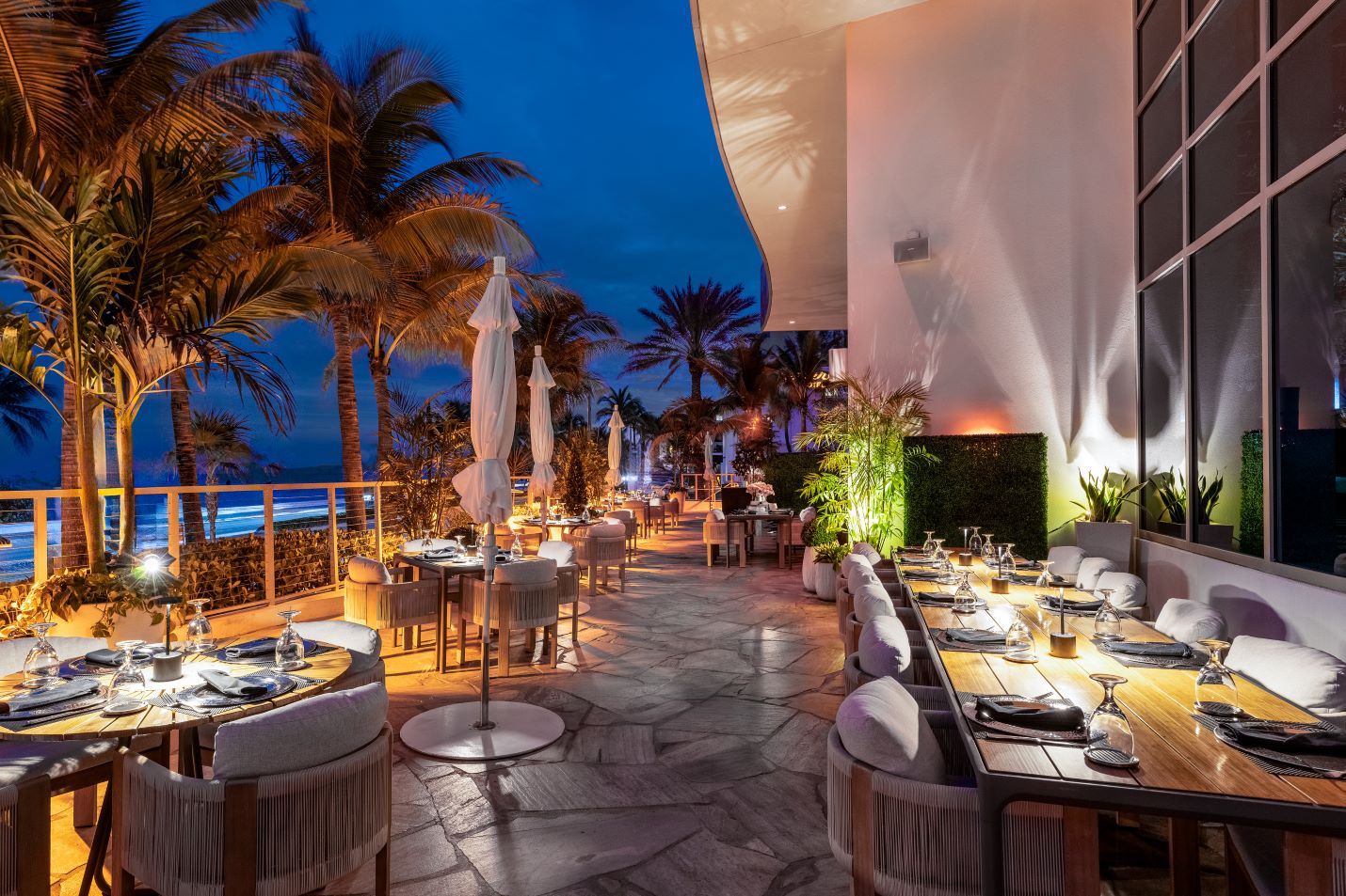 exterior of Truluck's Fort Lauderdale patio area with tables and cushioned chairs, palm trees and lots of up lighting.