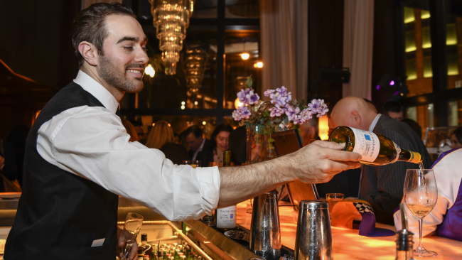 A bartender smiling and pouring a glass of white wine for a guest at the bartop
