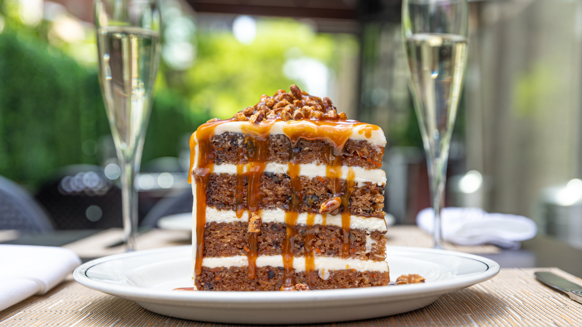 World class carrot cake on a plate drizzled in caramel with pecans on top and two glasses of champagne in the background