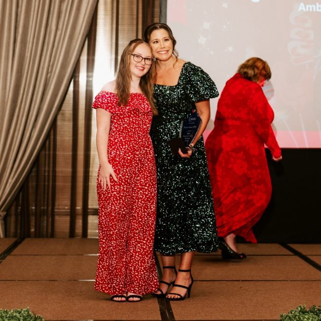 Amber in a green sequin dress and Sarah in a red dress on stage after Amber received an award