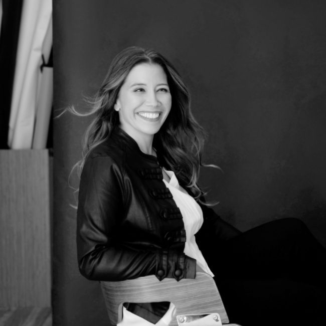 headshot of Amber sitting in a chair in a black leather jacket, smiling with hair slightly blowing left