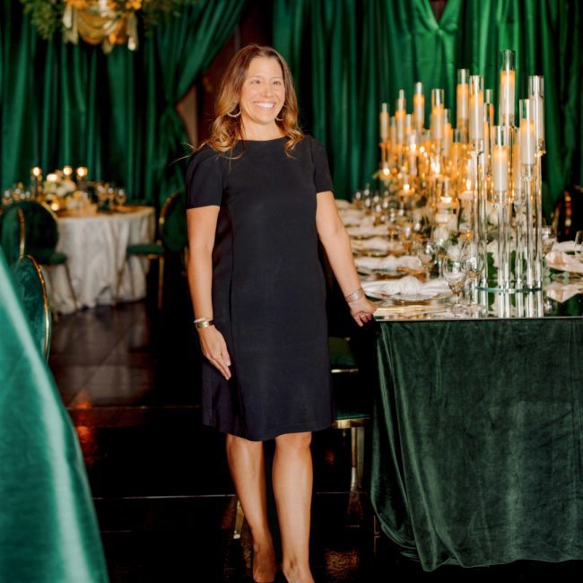 Amber in a black dress standing in front of a dining table dressed in emerald green linen with tall glass candles