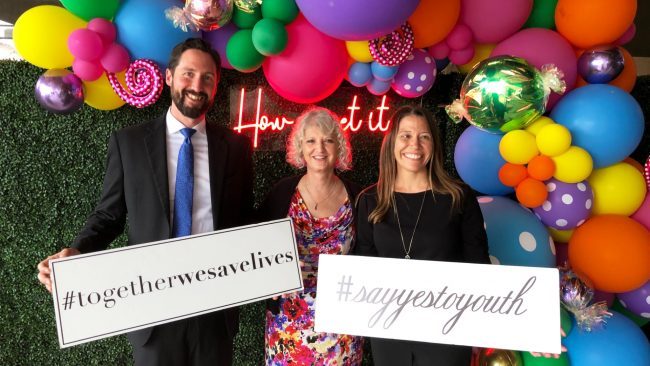 Chris and Amber with a partner from Yes to Youth in front of a colorful balloon backdrop with signs that say #together we save live and # say yes to youth