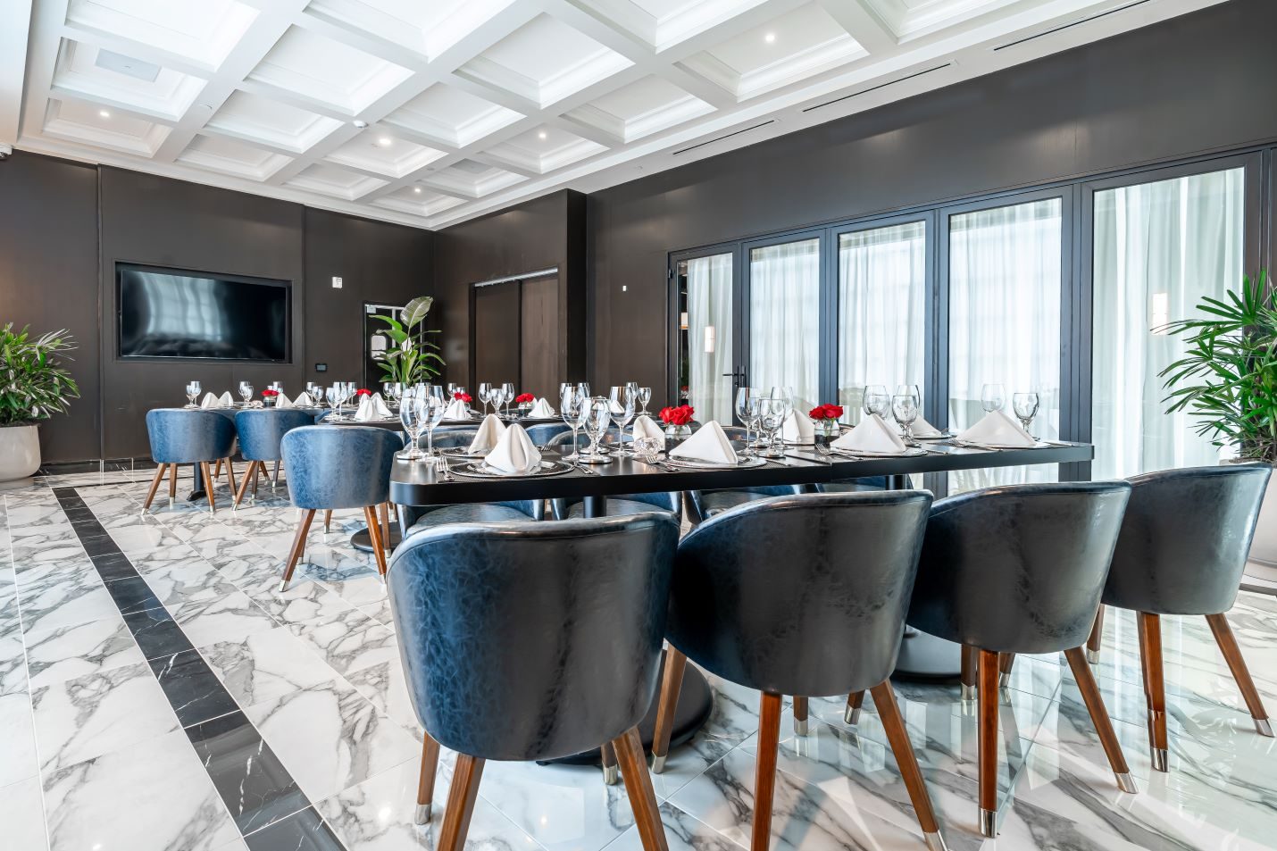private Trident room at Truluck's Plano setup with navy leather chairs, black table tops, white linen napkins, glassware, red roses and marble tile floors with white decorative ceiling, glassed doors and a large TV mounted on the wall