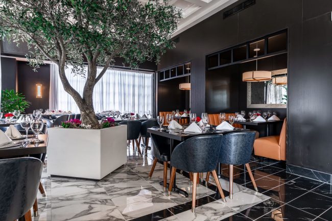 The Legacy group dining room with a large olive tree in a white planter, peacock blue chairs and black marble flooring