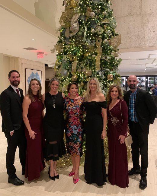 Amber and her colleagues all dressed up in evening attire in front of a tall Christmas tree decorated in gold.