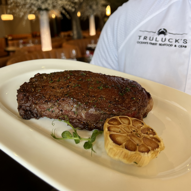 Truluck's chef coat in the background with a New York strip being held on a white plate with a half roasted garlic bulb and large floral tubes in the background