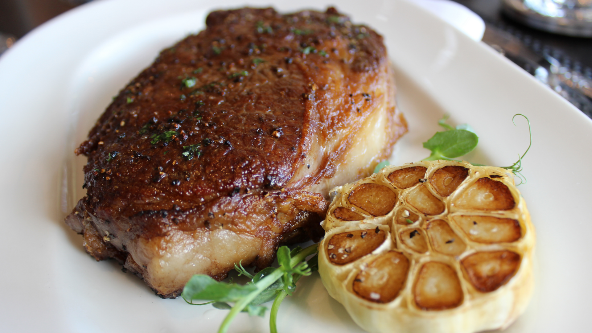 Ribeye steak on a white plate with a roasted garlic bulb and green parsley garnish