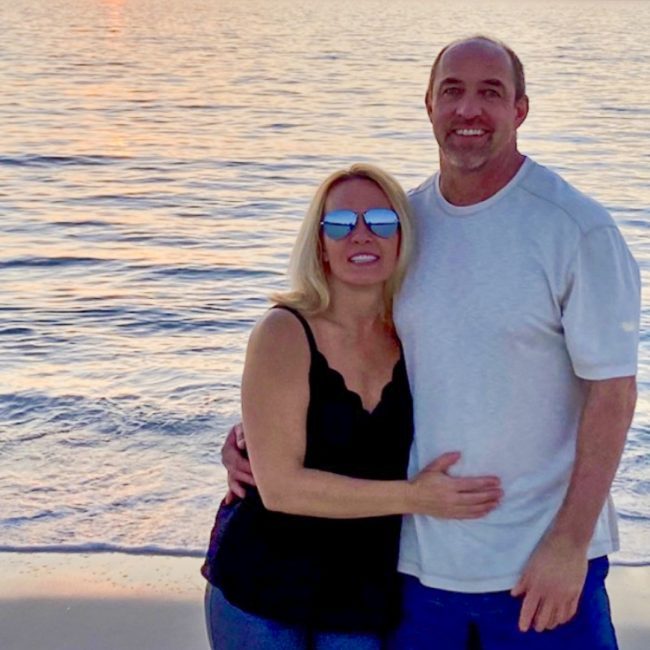 Ken and his wife in casual clothing in front of the ocean on the beach at sunset