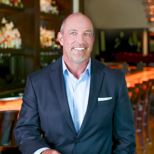 headshot of managing partner Ken Allen in a navy suit with the restaurant glowing bar in the background