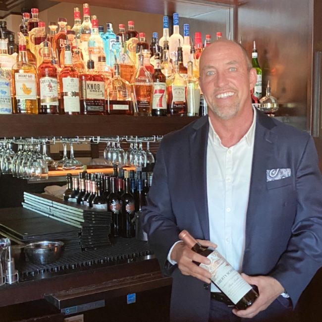 managing partner Ken Allen holding a bottle of wine while behind the bar in front of the back bar liquor