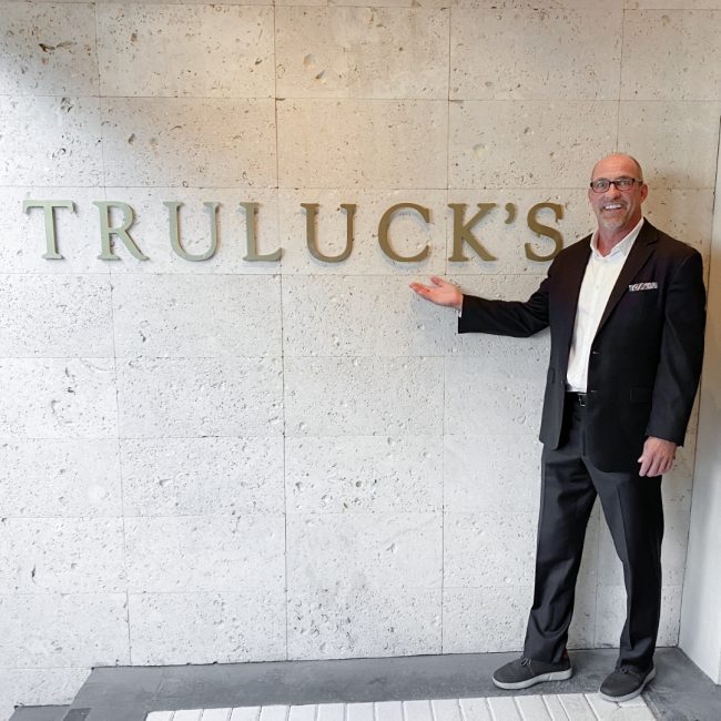 managing partner Ken Allen outside of the Naples location front entry dressed in a suit with his arm up under the Truluck's gold letters on the wall