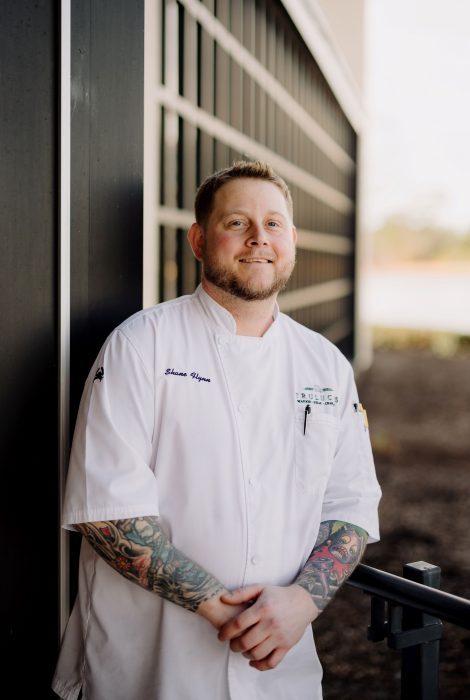 headshot of Chef Partner Shane Flynn in his Chef coat with his hands crossed standing outside of Truluck's Woodlands in front of the windows