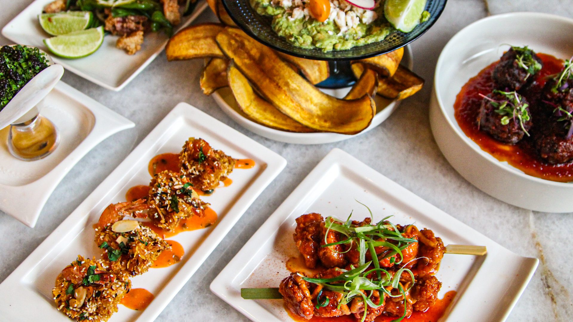 combination of happy hour food including hot and crunchy shrimp topped with slivered almond, hot honey chicken bites on skewers with shaved green onion, prime meatballs in house made steak sauce dressed with micro greens and blue crab guacamole with plantain chips
