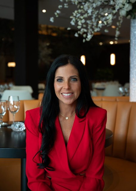 headshot of General manager Tambra Tatum in a red jacket in the main dining room with floral sprays in the background