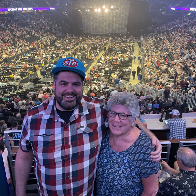 Keegan in a checked shirt and STP hat with his mom in a large arena to watch a concert