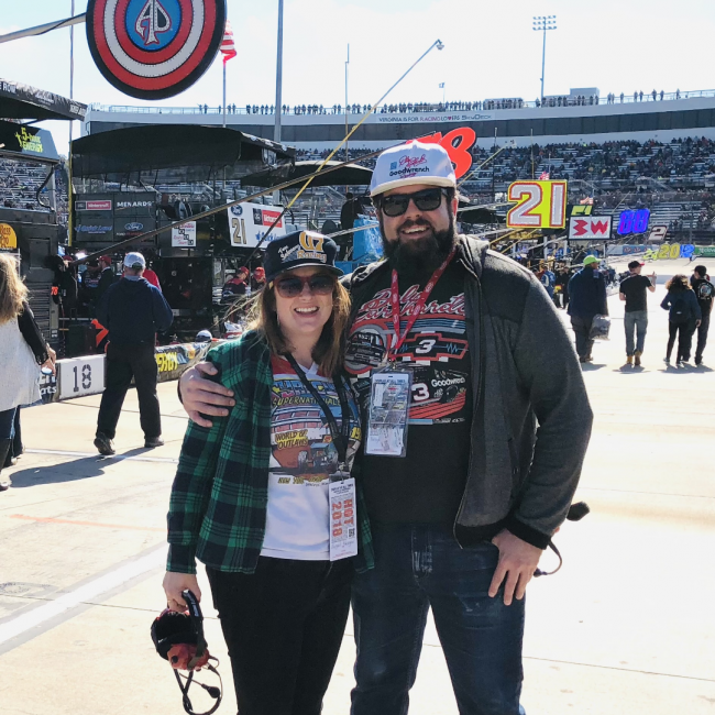 Keegan and his wife at an auto racing event standing down on the track in hats and sunglasses