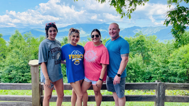 Jason with his wife and daughters outdoors inf front of a wooden post style fence and posing with a forest and mountainside in the background