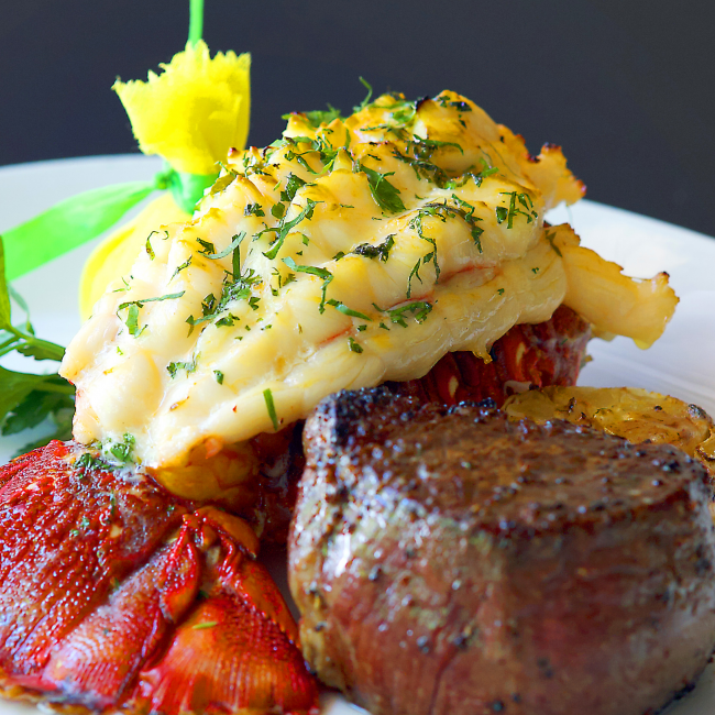 a whole lobster tail garnished with greenery and a filet steak with a lemon wrap paired on a white plate