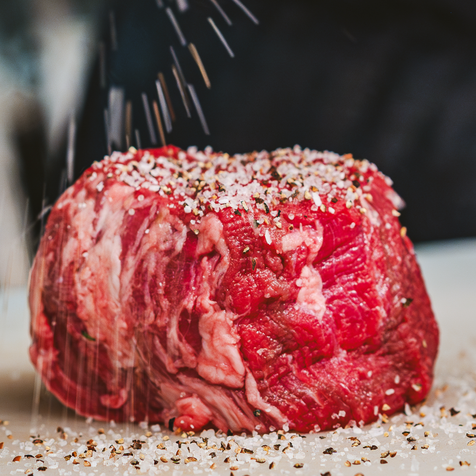 an uncooked 12 ounce filet of beef being dusted in salt and pepper in preparation for grilling