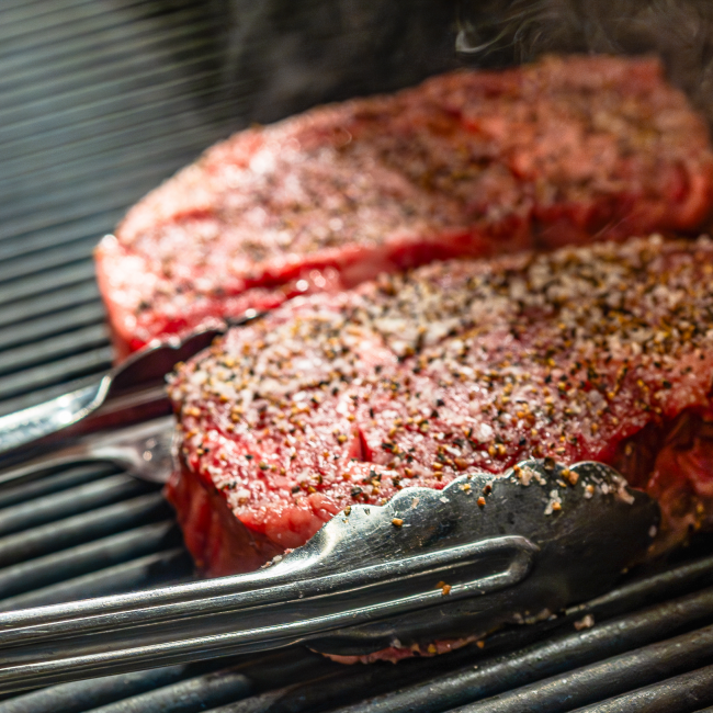 two new york strip steaks on a grill grate with steam coming off of them and tongs wrapped around one steak about to flip it over