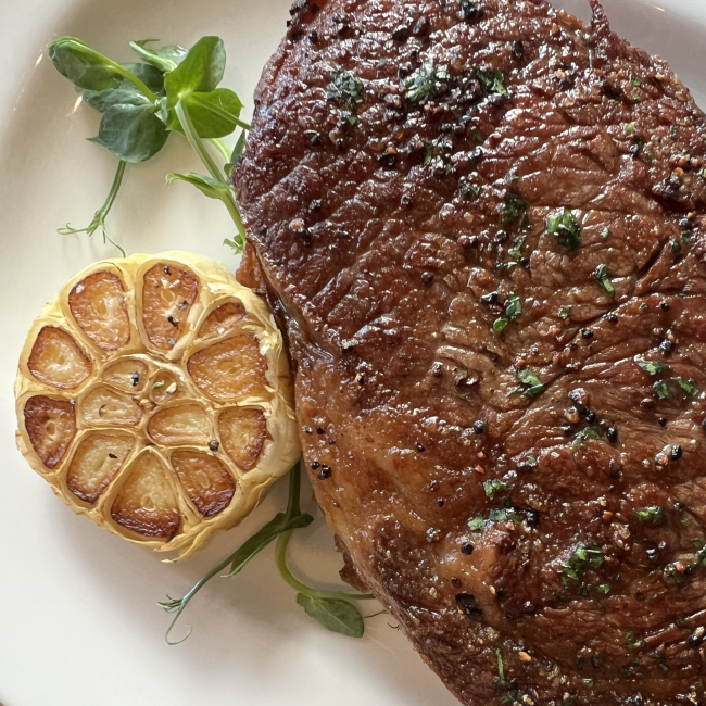 close up image of a ribeye steak crusted in pepper with a half clove of roasted garlic and fresh greenery