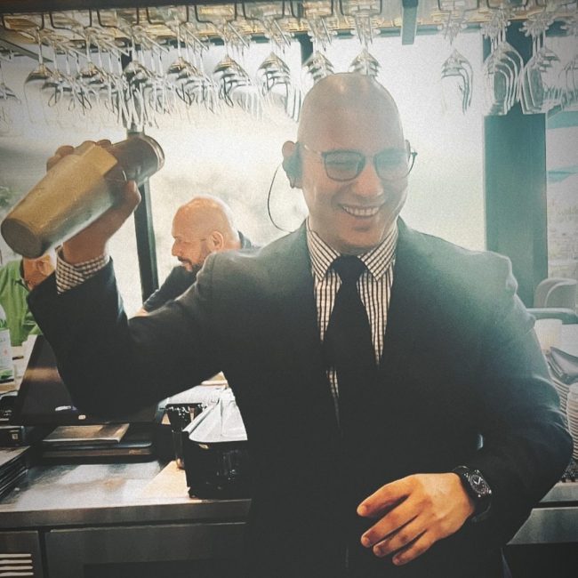 Miami beverage manager, Jesus Perez, in a suit and tie and glasses smiling and shaking up a cocktail in a shaker tin behind the bar