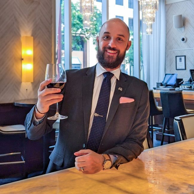 DC beverage manager Brian Dominic in a suite and tie raising a glass of red wine up at the bar top