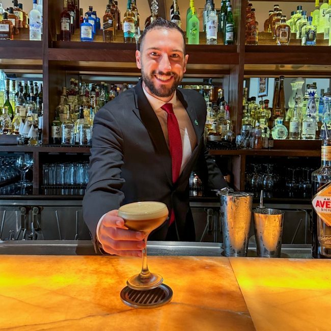 Arboretum Austin beverage manager, Cameron Stefanini, in a suite and tie behind the bar serving up a freshly made espresso martini