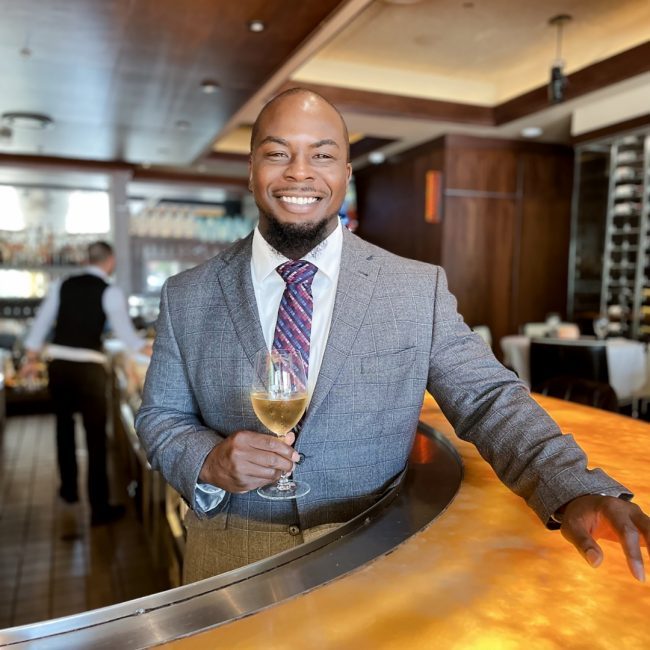 Dallas beverage manager, Deshon Ford, in a suit and tie smiling and poised in front of the bar top with a glass of white wine in hand