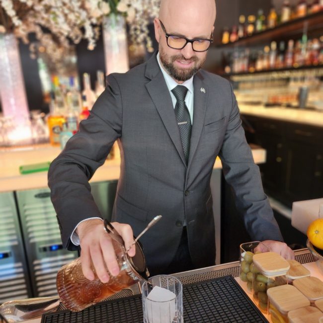 Plano beverage manager, Joshua Patrick, in a suit and tie behind the bar pouring a cocktail into a rocks glass filled with an ice block