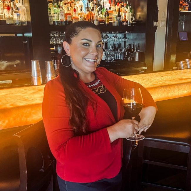 Naples beverage manager, Brooke Torrez in a red suit jacket smiling in front of the bar top and holding a glass of red wine