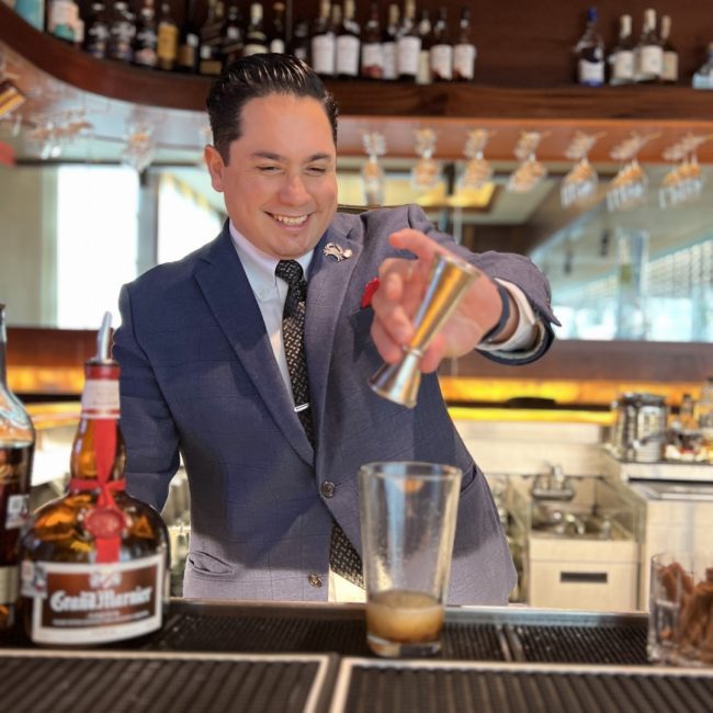 Woodlands beverage manager, Matthew Middleton, behind the bar in a suite and tie smiling and pouring Gran Marnier from a jigger into a pint glass while making a cocktail