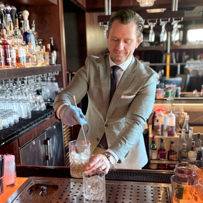 Rosemont beverage manager, David Talacha, behind the bar stirring a drink a glass cocktail pitcher.