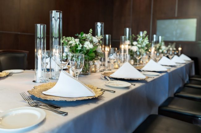 Naples room boardroom style table setting with gold chargers, white napkins, cylinder glass centerpieces with fresh flowers and candles