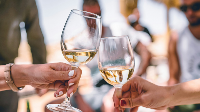 two women's hands holding white wine glasses and clinking them together in a toast fashion with a few people blurred in the background