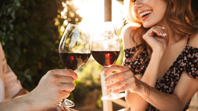 A smiling lady and mane toasting red wine glasses with sunlight shining in the background