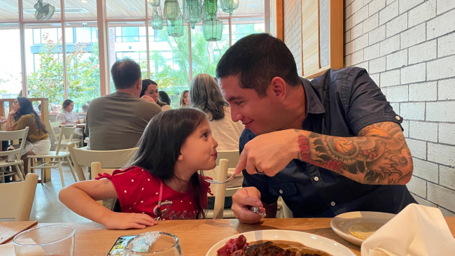 Fern and his daughter looking at each other face to face in a restaurant and smiling.