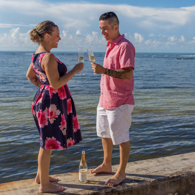 fern and his wife holding two glasses of champagne and toasting in front of the ocean