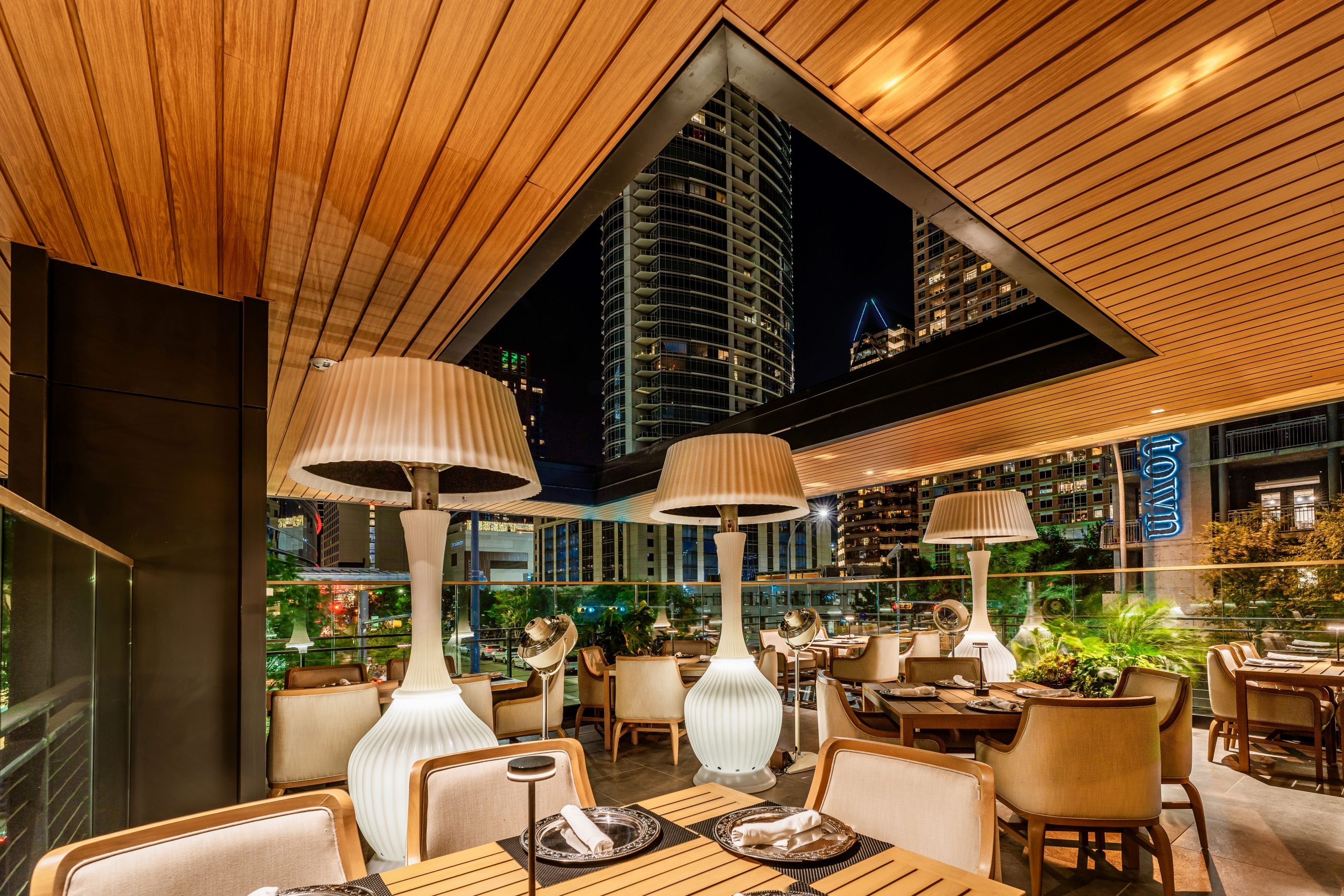 Truluck's Austin downtown outdoor patio at night time set with teakwood tables and cushioned wooden resort style chairs and heater style lamp lights. There is a view of the Austonian and the JW Marriott through the ceiling cutout.