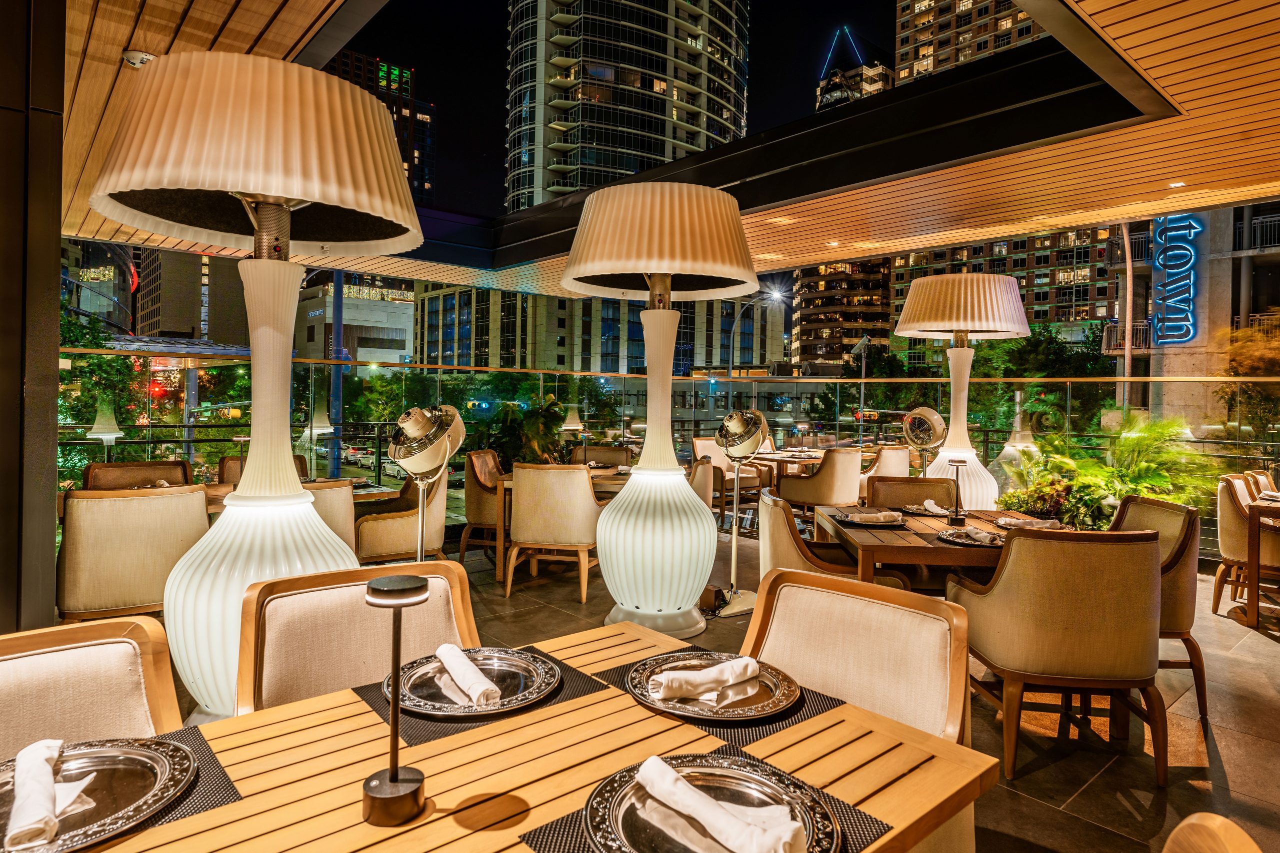 Truluck's Austin downtown outdoor patio at night time set with teakwood tables and cushioned wooden resort style chairs and heater style lamp lights. There are skyline views of downtown through the cutout.