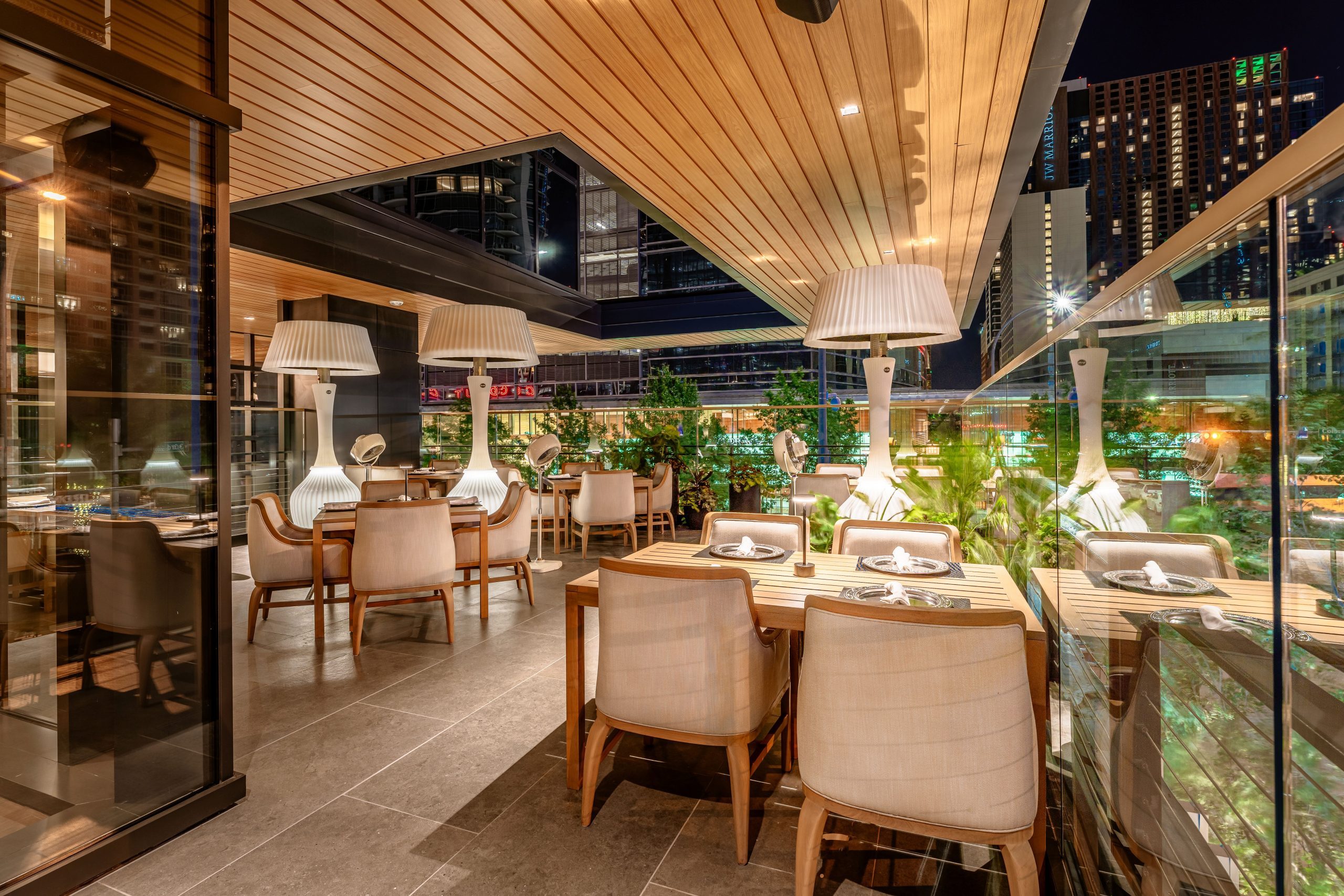 Truluck's Austin downtown outdoor patio at night time set with teakwood tables and cushioned wooden resort style chairs and heater style lamp lights.