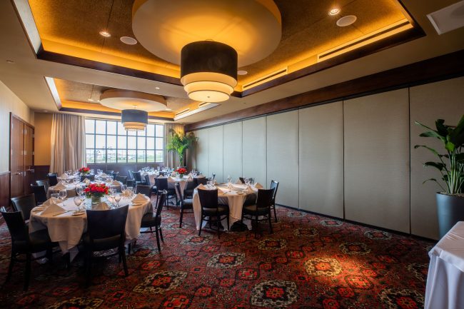 Truluck's Rosemont Pearl Room view from the front entry of the room. The room is set with round tables of 6 dressed in white linen. There is large window with white drapes at the far end of the room.