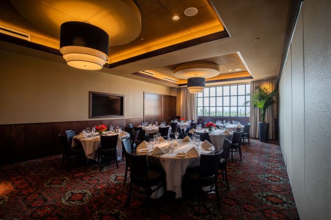 Truluck's Rosemont Pearl room view from the front corner of the room looking toward the window on the far wall. There is a TV on the wall and the room is set up with round tables and chairs.