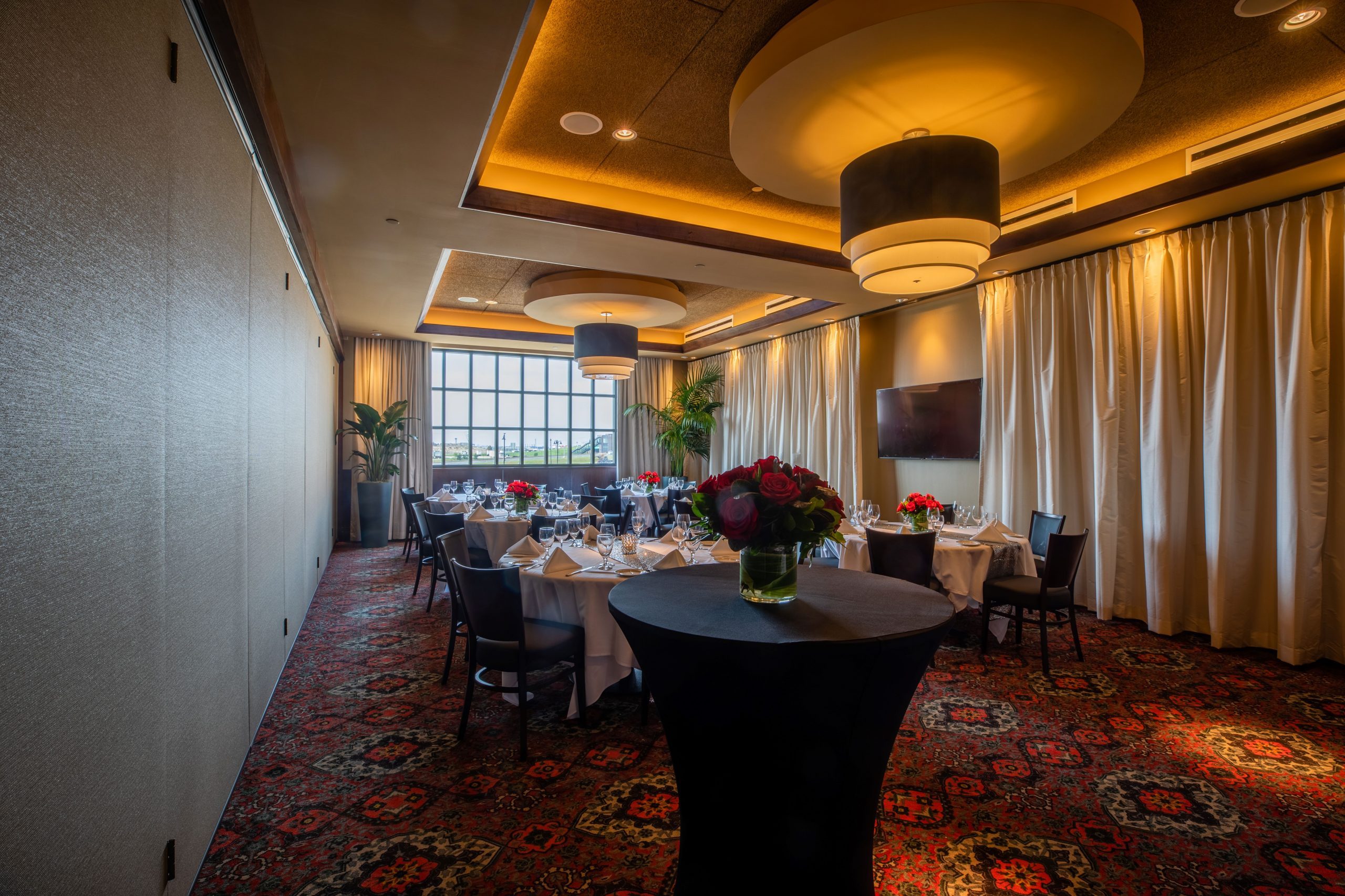 Truluck's Rosemont Naples room view from the far left front corner. The room is setup with a tall cocktail table dressed in black linen in the front and 8 top round tables with chairs dressed in white linen are the background. There is a large window on the far wall with curtain.