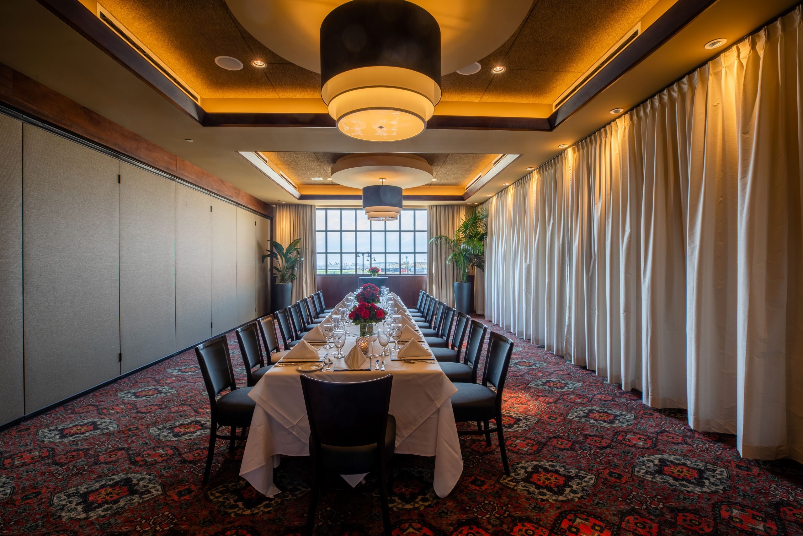 Truluck's Rosemont Naples room view from the entry door. The room is setup with a boardroom style table dressed in white linen with red rose floral centerpieces. The left wall is the airwall that divides the stone crab room into two smaller room. There is a window at the far end with curtains.