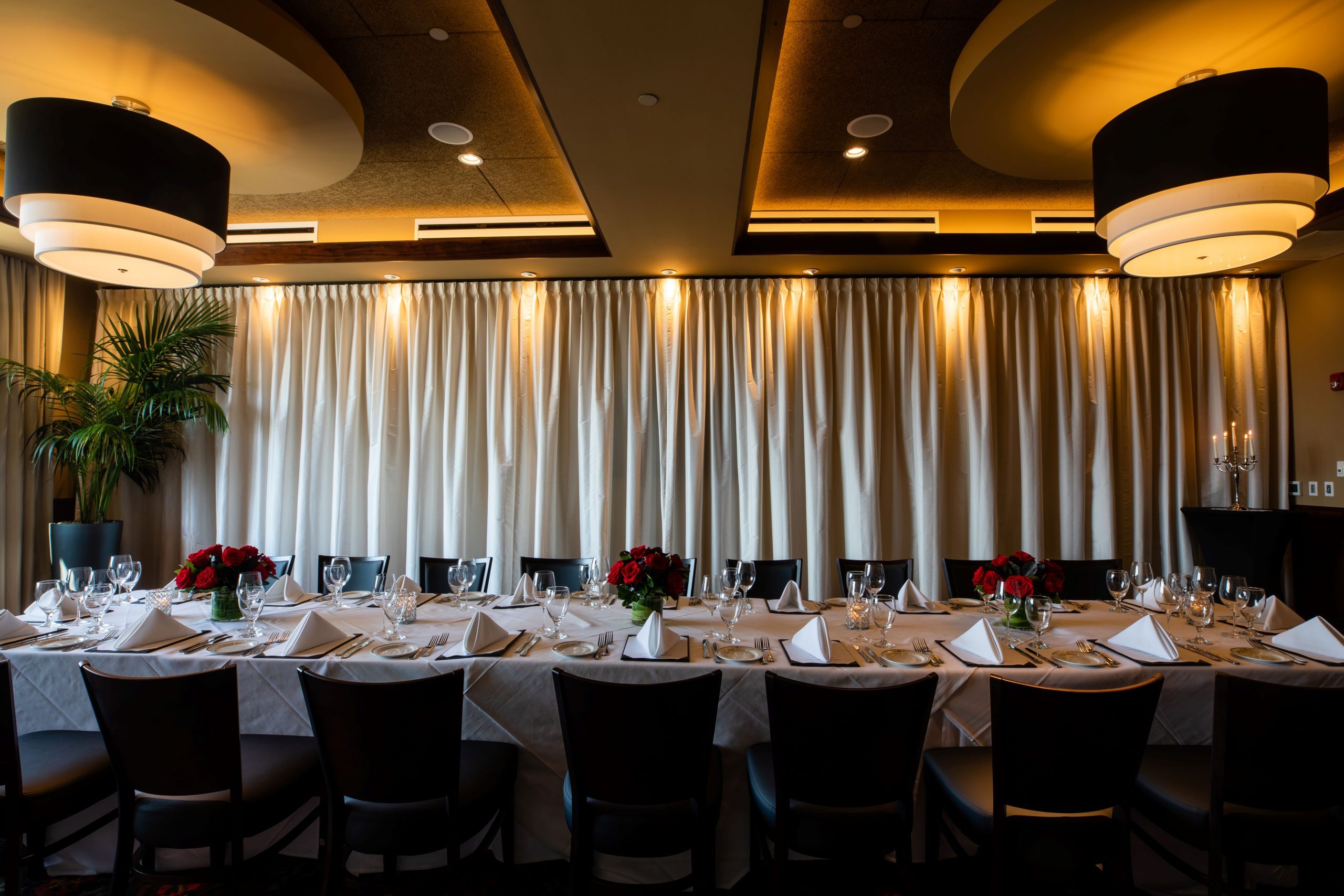 Truluck's Rosemont Naples room setup with a boardroom style table dressed in white linen with red roses on the tabletop. The back wall is dressed in a white curtain with illumination.