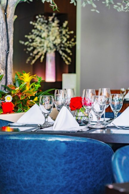 A close up shot of a mezzanine table at the Truluck's Austin downtown location. The table is set with blue leather chairs, red rose centerpieces and glassware with white linen napkins. There is a large floral in the background.