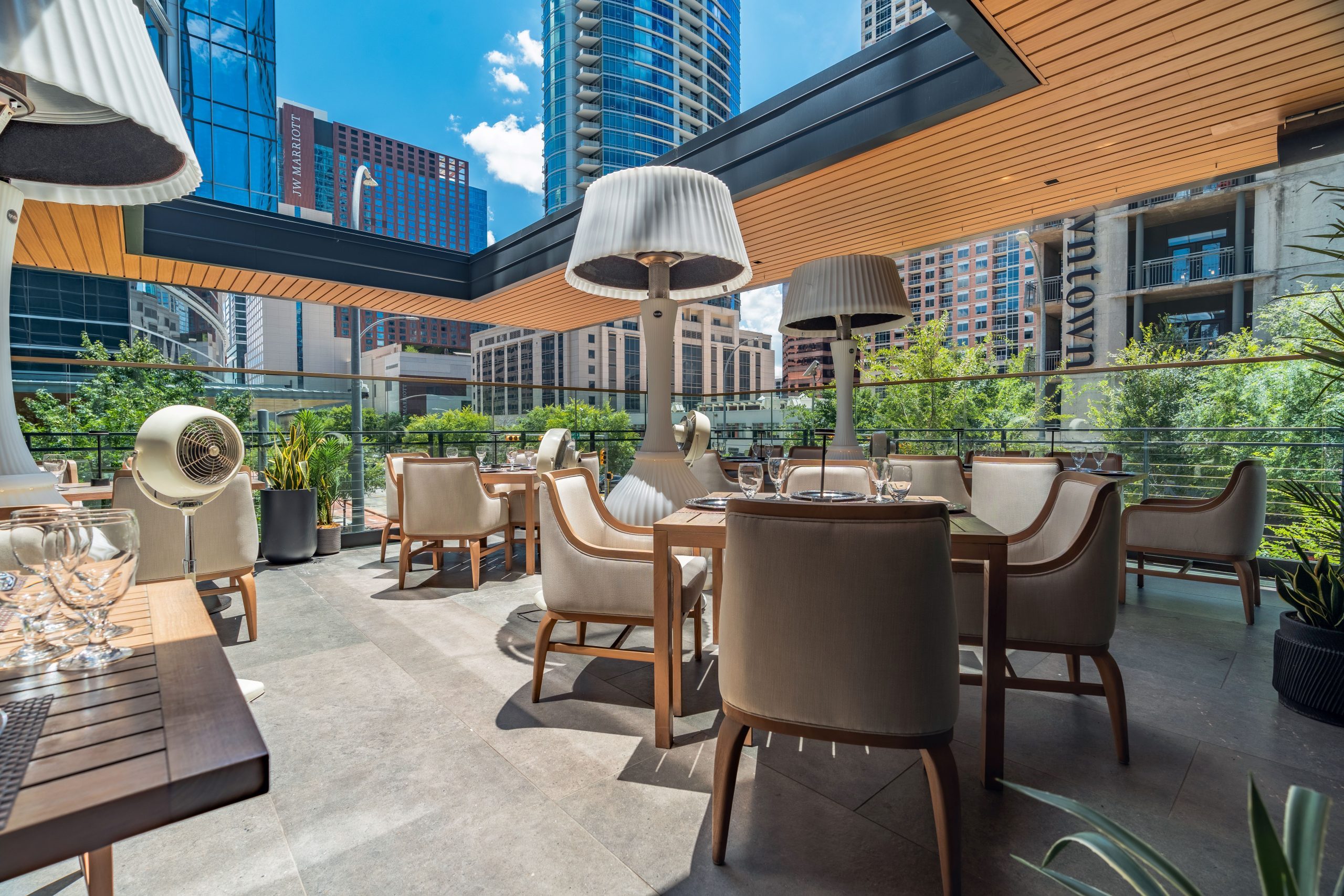 Truluck's Austin downtown outdoor patio during the daytime set with teakwood tables and cushioned wooden resort style chairs. The ceiling has a large cutout with skyline views of downtown and the JW Marriott and Austonian in the distance.