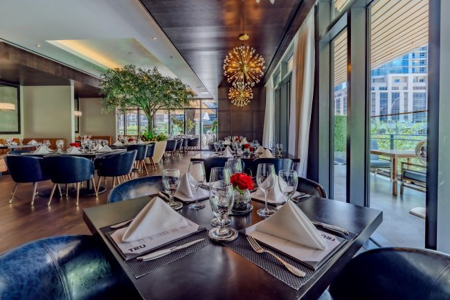 Truluck's Austin downtown upstairs mezzanine garden room view from the west end of the room. There is a large olive tree in the center of the room. The tables are wood with navy leather chairs and there are gold sputnik style chandeliers hanging over them.